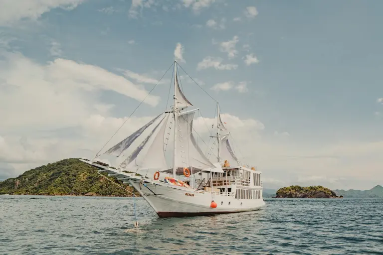 Phinisi Akassa berlambung putih dengan layar terkembang di dekat pulau hijau Taman Nasional Komodo