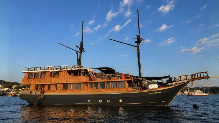 Kapal phinisi kayu Budi Utama berlabuh di perairan Labuan Bajo, lambung gelap dan dek kayu bertingkat