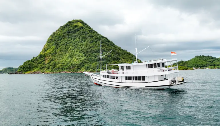 The white wooden Deden Putra at anchor off Labuan Bajo with green hills behind