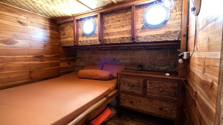 Kabin ranjang queen Sabbar Cruise berdinding kayu dengan dua jendela bulat dan laci kayu