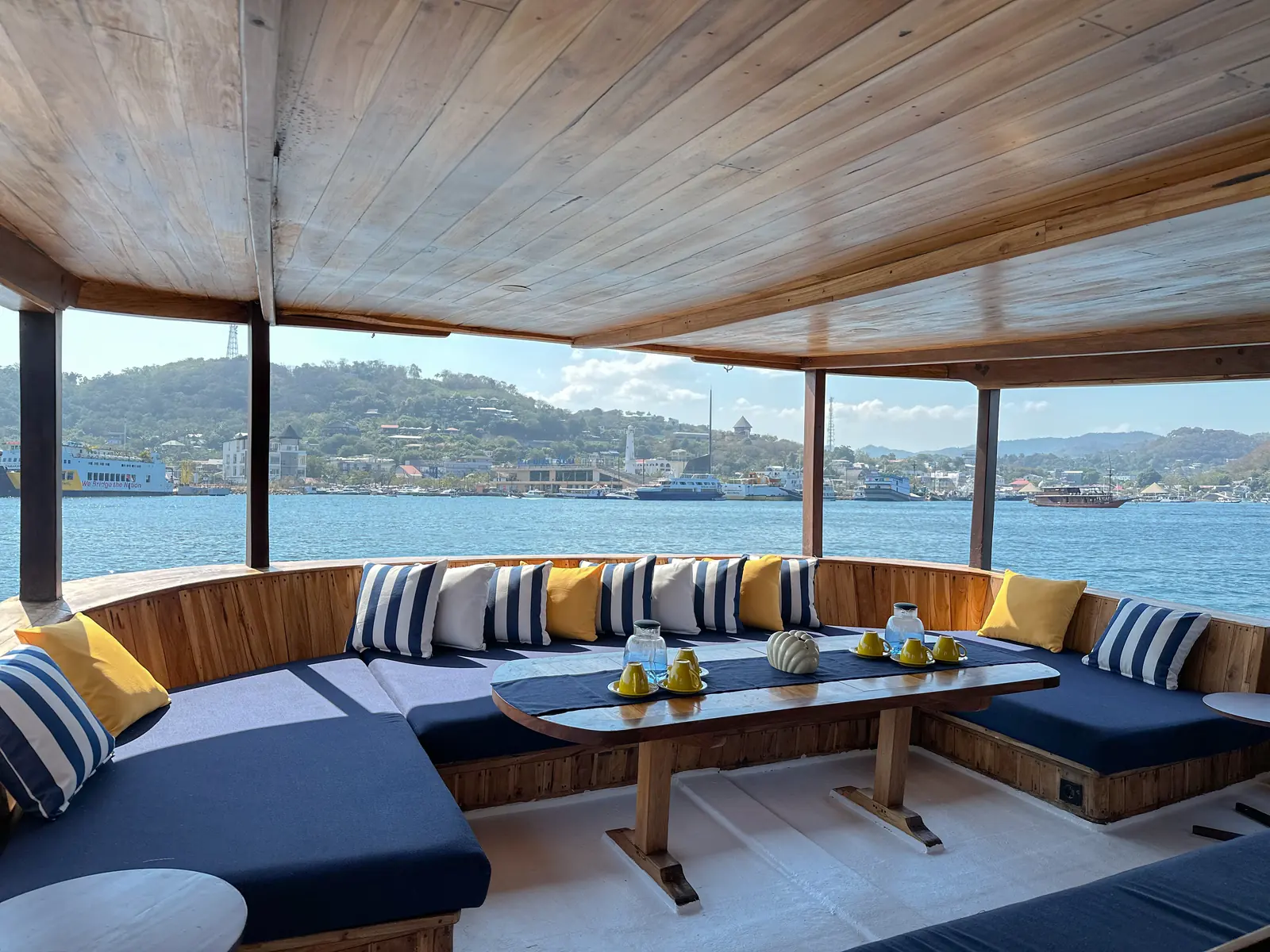 Sofa biru berbantal garis di dek beratap Vhalé menghadap pelabuhan Labuan Bajo